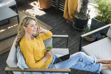 Portrait of smiling mature woman sitting on terrace with digital tablet and glass of smoothie - UUF20541