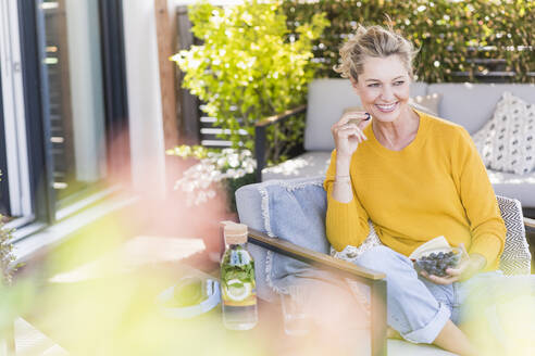 Porträt einer glücklichen reifen Frau, die auf der Terrasse sitzt und Blaubeeren isst - UUF20543