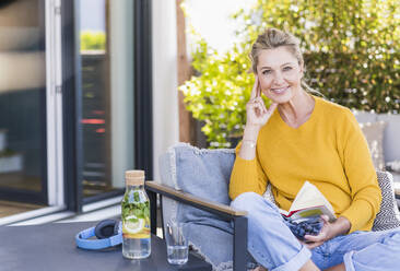 Porträt einer lächelnden reifen Frau, die mit einer Schüssel Blaubeeren und einem Buch auf der Terrasse sitzt - UUF20548