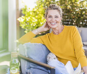 Porträt einer glücklichen reifen Frau, die mit einem Buch auf der Terrasse sitzt - UUF20552