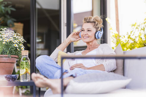 Porträt einer lächelnden reifen Frau, die auf einer Terrasse sitzt und mit Kopfhörern Musik hört - UUF20553