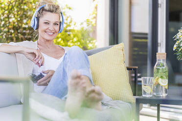 Porträt einer lächelnden reifen Frau, die auf einer Terrasse sitzt und mit Kopfhörern Musik hört, während sie Blaubeeren isst - UUF20556