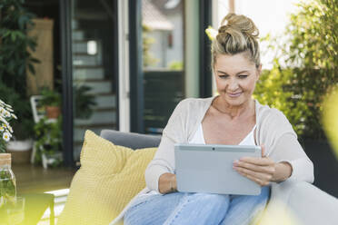 Porträt einer lächelnden reifen Frau, die auf einer Terrasse sitzt und auf ein digitales Tablet schaut - UUF20557