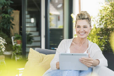 Portrait of happy mature woman sitting on terrace with digital tablet - UUF20558