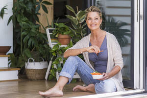 Porträt einer lächelnden reifen Frau, die bei offener Terrassentür auf dem Boden sitzt und Fingerfood isst - UUF20564