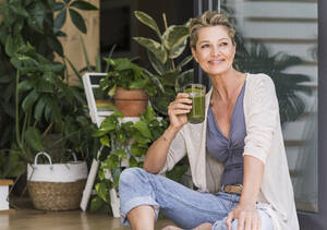 Porträt einer entspannten reifen Frau, die an der offenen Terrassentür mit einem Glas grünem Smoothie sitzt - UUF20565