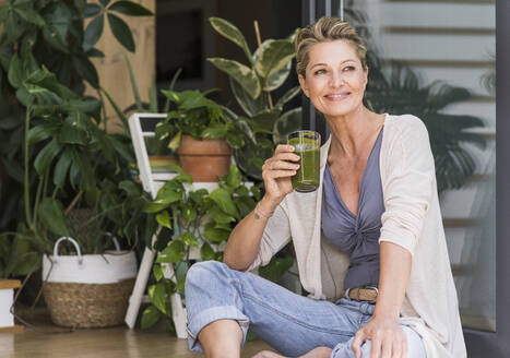 Portrait of relaxed mature woman sitting at open terrace door with glass of green smoothie - UUF20565