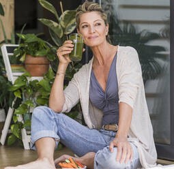 Portrait of smiling mature woman sitting at open terrace door drinking green smoothie - UUF20567