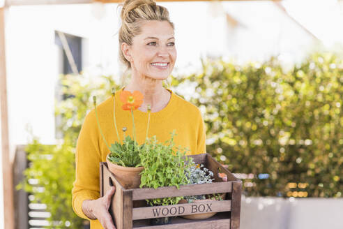 Porträt einer glücklichen reifen Frau mit Holzkiste mit Pflanzen auf der Terrasse - UUF20580