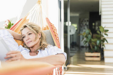 Porträt einer reifen Frau, die sich in einer Hängematte auf einer Terrasse entspannt und ein digitales Tablet benutzt - UUF20592