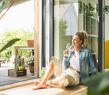 Porträt einer lächelnden reifen Frau, die bei offener Terrassentür auf dem Boden sitzt und ein Glas Wasser trinkt - UUF20596