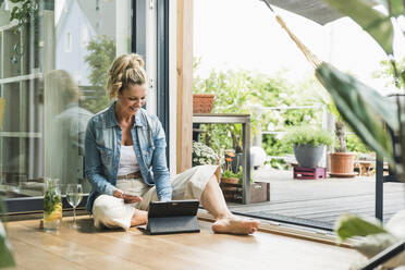 Lächelnde reife Frau, die bei offener Terrassentür auf dem Boden sitzt und ein Smartphone und ein digitales Tablet benutzt - UUF20598