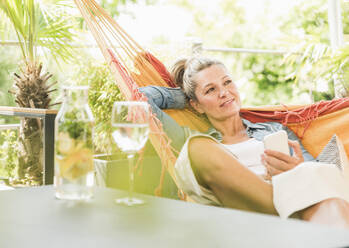 Porträt einer nachdenklichen reifen Frau mit Smartphone, die sich in einer Hängematte auf einer Terrasse entspannt - UUF20600