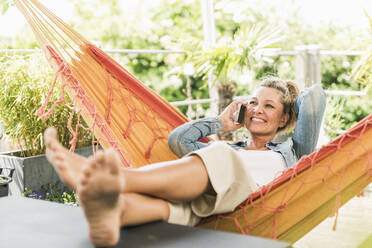 Portrait of happy mature woman on the phone relaxing in hammock on terrace - UUF20603