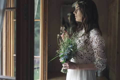 Junge Frau in elegantem Hochzeitskleid mit Blumenstrauß, die aus dem Fenster schaut, lizenzfreies Stockfoto