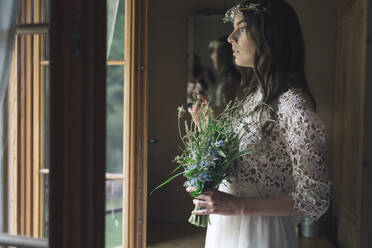 Junge Frau in elegantem Hochzeitskleid mit Blumenstrauß, die aus dem Fenster schaut - ALBF01260