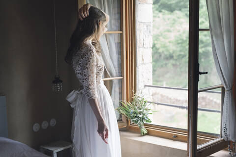 Junge Frau in elegantem Hochzeitskleid mit Blumenstrauß, die aus dem Fenster schaut, lizenzfreies Stockfoto