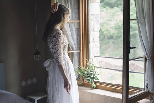 Young woman in elegant wedding dress holding bouquet looking out of the window - ALBF01264