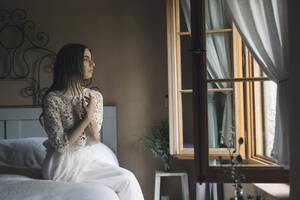 Young woman in elegant wedding dress sitting on bed looking out of the window - ALBF01266
