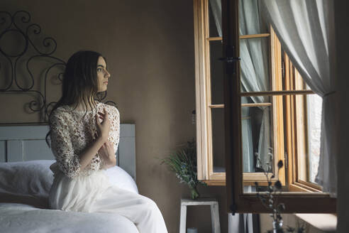 Junge Frau in elegantem Hochzeitskleid sitzt auf dem Bett und schaut aus dem Fenster - ALBF01266