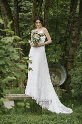 Junge Frau in elegantem Hochzeitskleid mit Blumenstrauß auf einer Bank stehend - ALBF01269