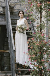Junge Frau in elegantem Hochzeitskleid mit Blumenstrauß auf einer Treppe stehend - ALBF01282
