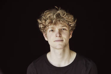 Portrait of young man with curly blond hair against black background - FMKF06168