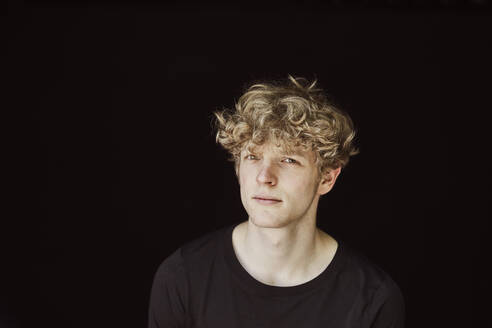 Porträt eines skeptischen jungen Mannes mit lockigem blondem Haar vor schwarzem Hintergrund - FMKF06169