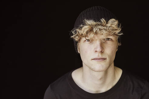 Portrait of young man with curly blond hair wearing black cap against black background - FMKF06171