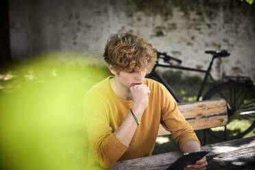 Junger Mann sitzt am Biertisch im Garten und benutzt ein digitales Tablet - FMKF06174