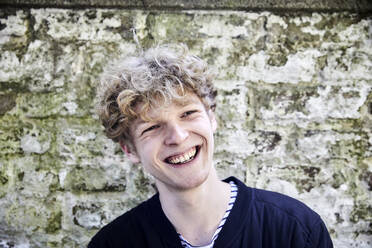 Porträt eines lachenden jungen Mannes mit lockigem blondem Haar vor einer verwitterten Wand - FMKF06189