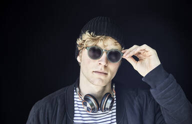 Porträt eines jungen Mannes mit Kopfhörern, Mütze und Sonnenbrille vor schwarzem Hintergrund - FMKF06197
