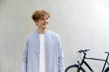 Portrait of smiling young man with curly blond hair standing in front of grey wall - FMKF06201