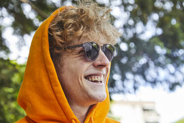 Portrait of laughing young man wearing sunglasses and orange hood - FMKF06204