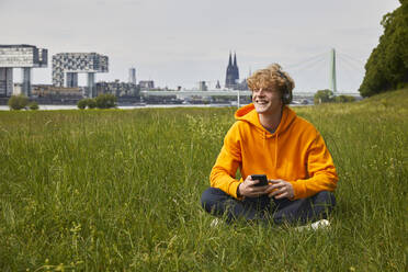 Happy young man sitting on a meadow listening music with headphones, Cologne, Germany - FMKF06206