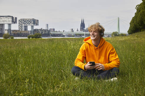 Glücklicher junger Mann, der auf einer Wiese sitzt und mit Kopfhörern Musik hört, Köln, Deutschland - FMKF06206