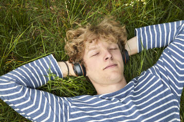 Portrait of young man with curly blond hair lying on a meadow listening music with headphones - FMKF06207