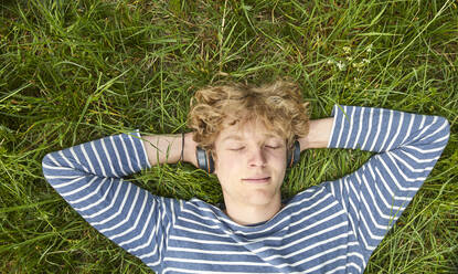 Portrait of young man with curly blond hair lying on a meadow listening music with headphones - FMKF06208