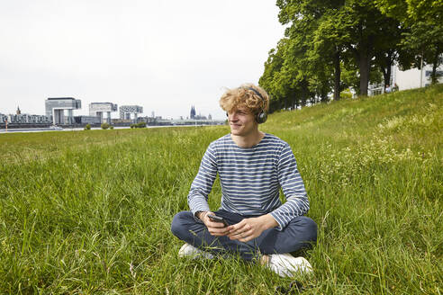 Smiling young man sitting on a meadow listening music with headphones - FMKF06210