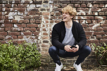 Portrait of laughing young man with smartphone chrouching in front of brick wall - FMKF06212