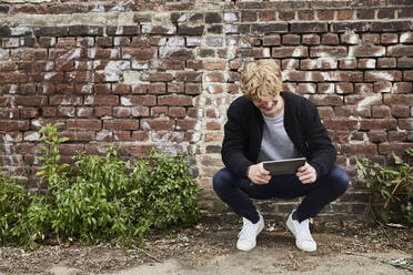 Lächelnder junger Mann, der vor einer Backsteinmauer hockt und auf ein digitales Tablet schaut - FMKF06213