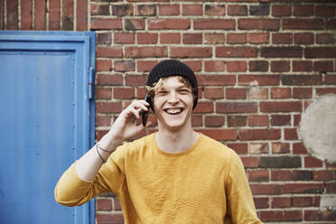 Porträt eines lachenden jungen Mannes am Telefon vor einer Backsteinmauer - FMKF06218