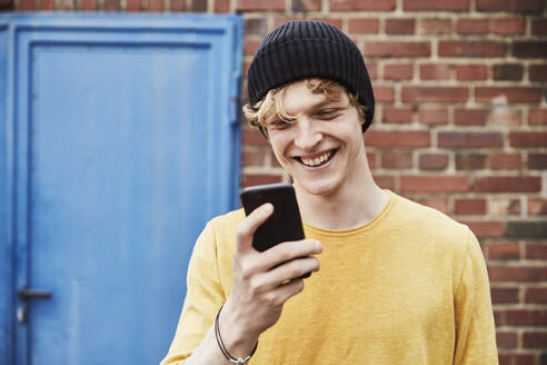 Porträt eines glücklichen jungen Mannes mit Mütze, der vor einer Backsteinmauer auf sein Smartphone schaut - FMKF06219