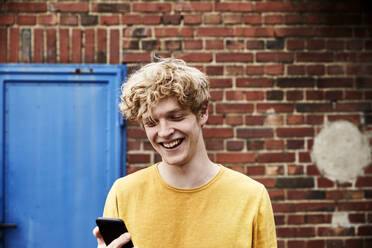 Porträt eines lachenden jungen Mannes, der vor einer Backsteinmauer auf sein Smartphone schaut - FMKF06220