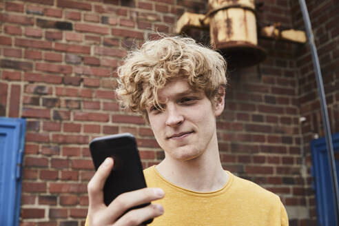 Porträt eines lächelnden jungen Mannes mit lockigem blondem Haar, der im Freien auf sein Smartphone schaut - FMKF06222