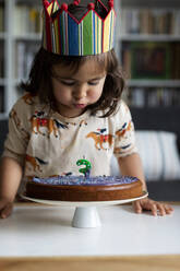 Porträt eines kleinen Mädchens, das zu Hause eine Kerze auf ihrem Geburtstagskuchen ausbläst - VABF03025