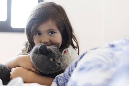 Porträt eines kleinen Mädchens, das eine graue Katze auf dem Bett kuschelt - VABF03031