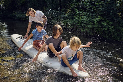 Father pushing boys splashing water while sitting on paddleboard in stream - PWF00115