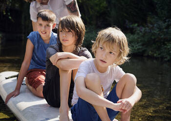 Thoughtful boys sitting on paddleboard in stream at forest - PWF00116