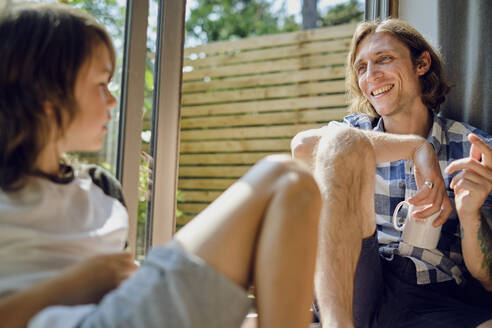 Happy father talking to son while sitting by window at home - PWF00124